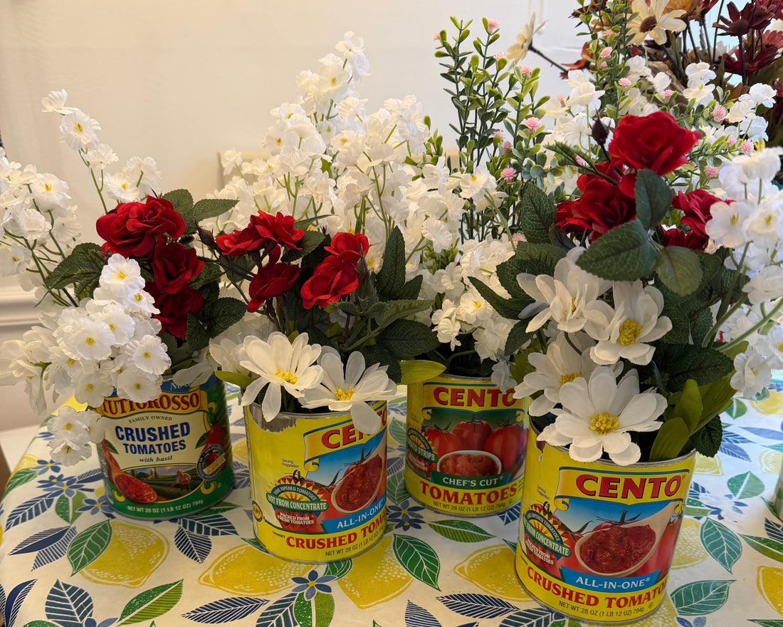 Four Italian tomato cans — Cento and Tuttorosso — filled with red roses, white daisies, and greenery, styled as party centerpieces on a lemon-print tablecloth for a Ciao Baby Italian-themed baby shower