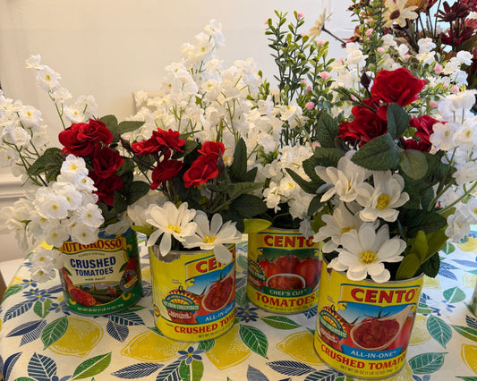 Four Italian tomato cans — Cento and Tuttorosso — filled with red roses, white daisies, and greenery, styled as party centerpieces on a lemon-print tablecloth for a Ciao Baby Italian-themed baby shower