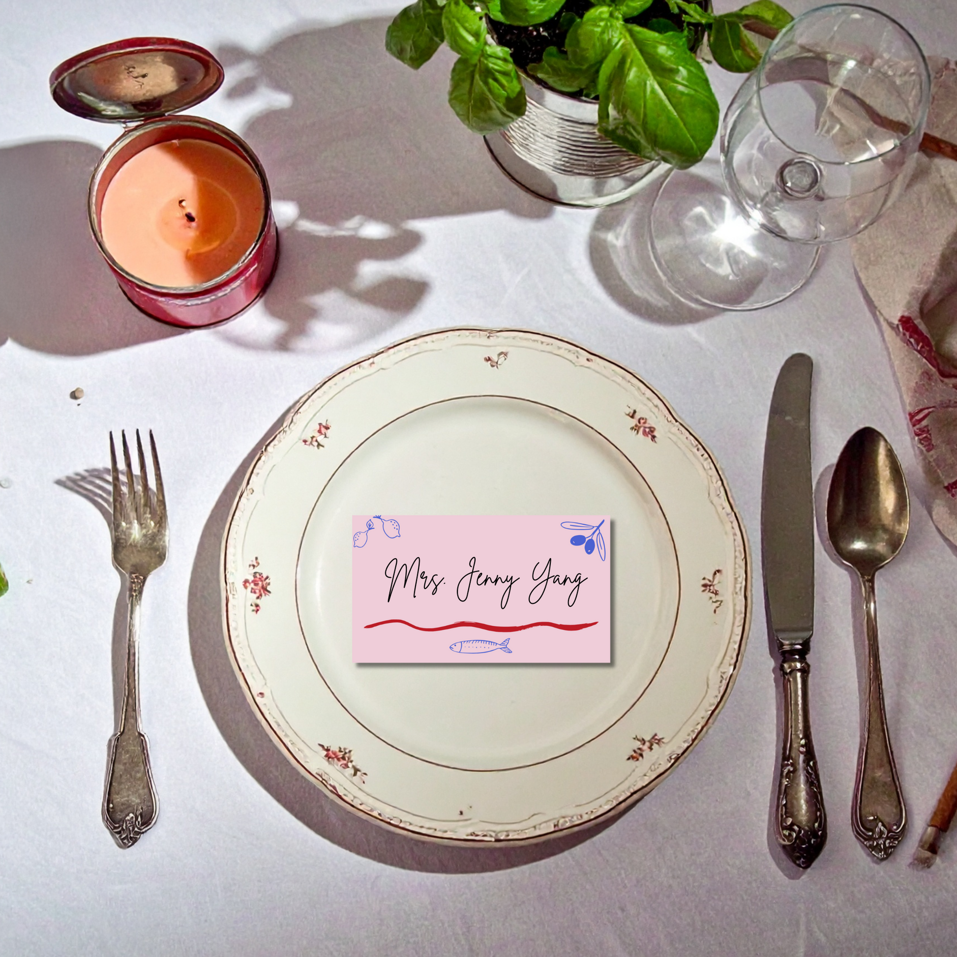 Dining table setting with a plate, cutlery, and a pink By Melon place card on a white tablecloth.