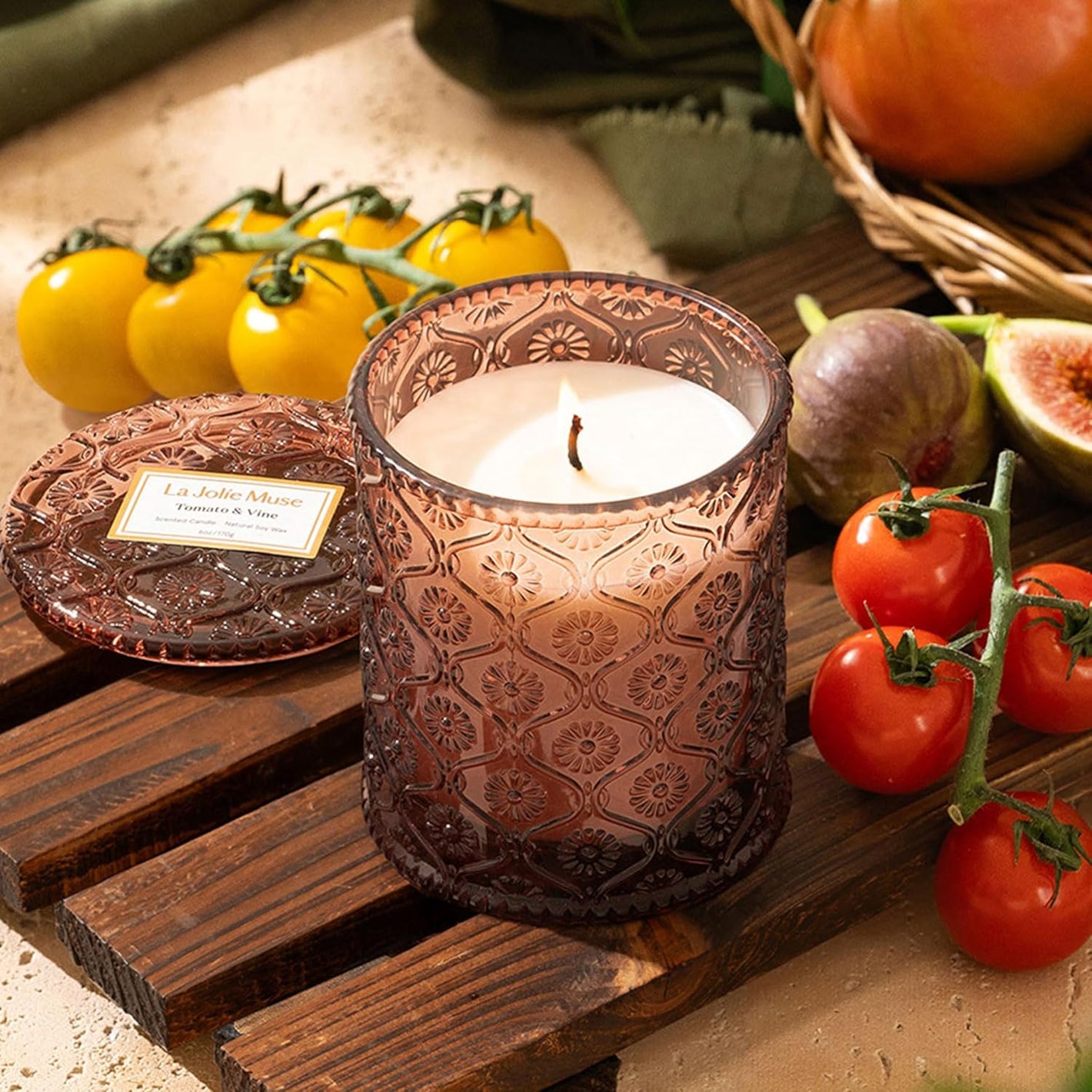 Decorative candle in a textured holder on a wooden surface with tomatoes and figs.